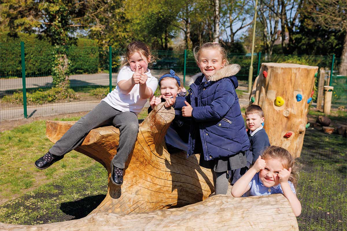 Colour photo of young children sitting on natural wooden log, smiling and putting their thumbs up to the camera in a school playground