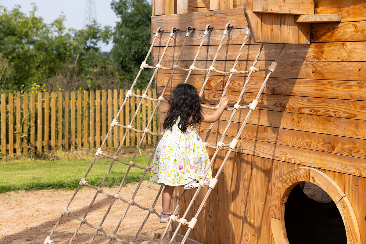 Colour photo of young girl climbing up net against wooden playground structure