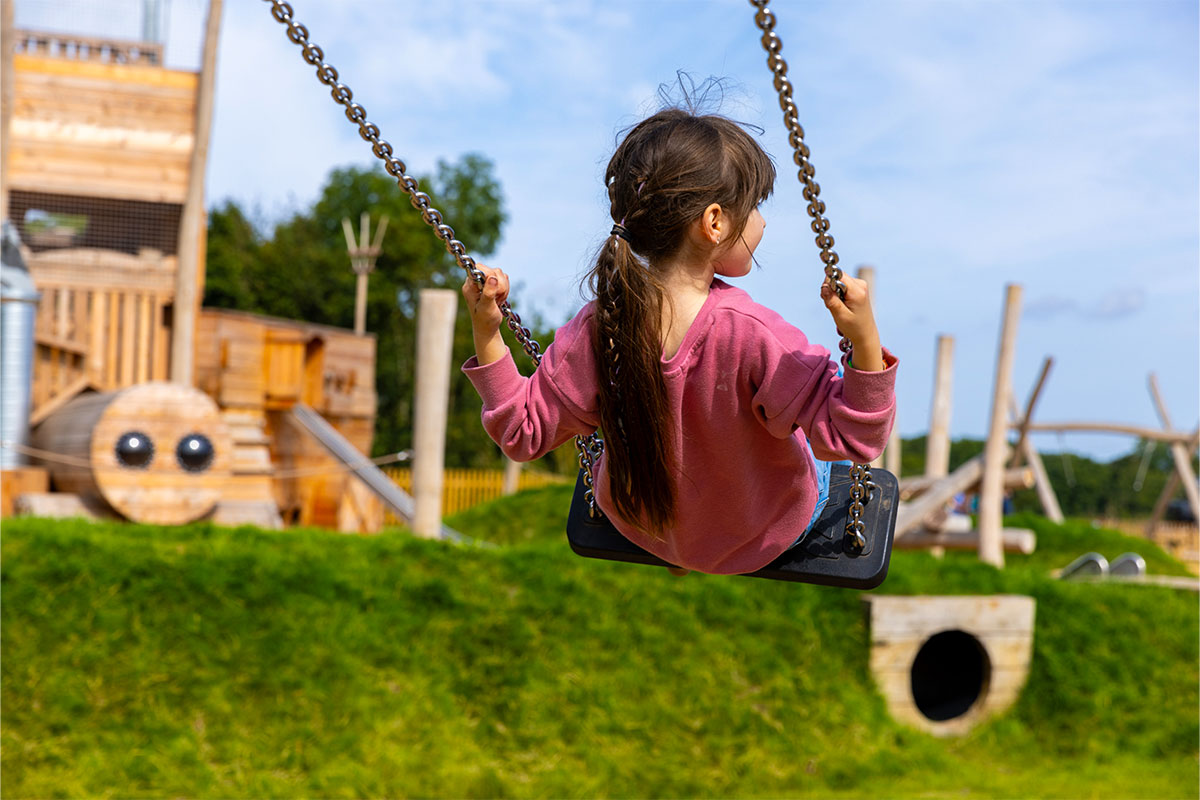 Colour photo of young girl on swing with Churchfields Farm adventure playground in the background