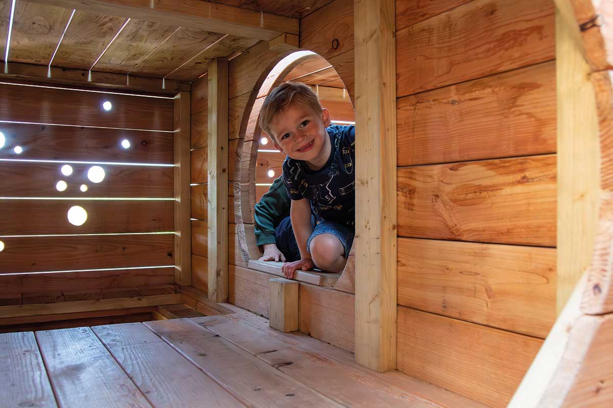 Colour photo of child crouching and smiling in hidey hole in wooden Festyland theme park playground