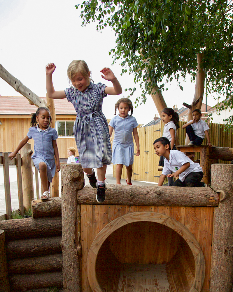 Children playing on a wooden playground structure in school uniforms. Their faces are happy. The playground is set against a backdrop of a fence, greenery, and a cloudy but bright sky.