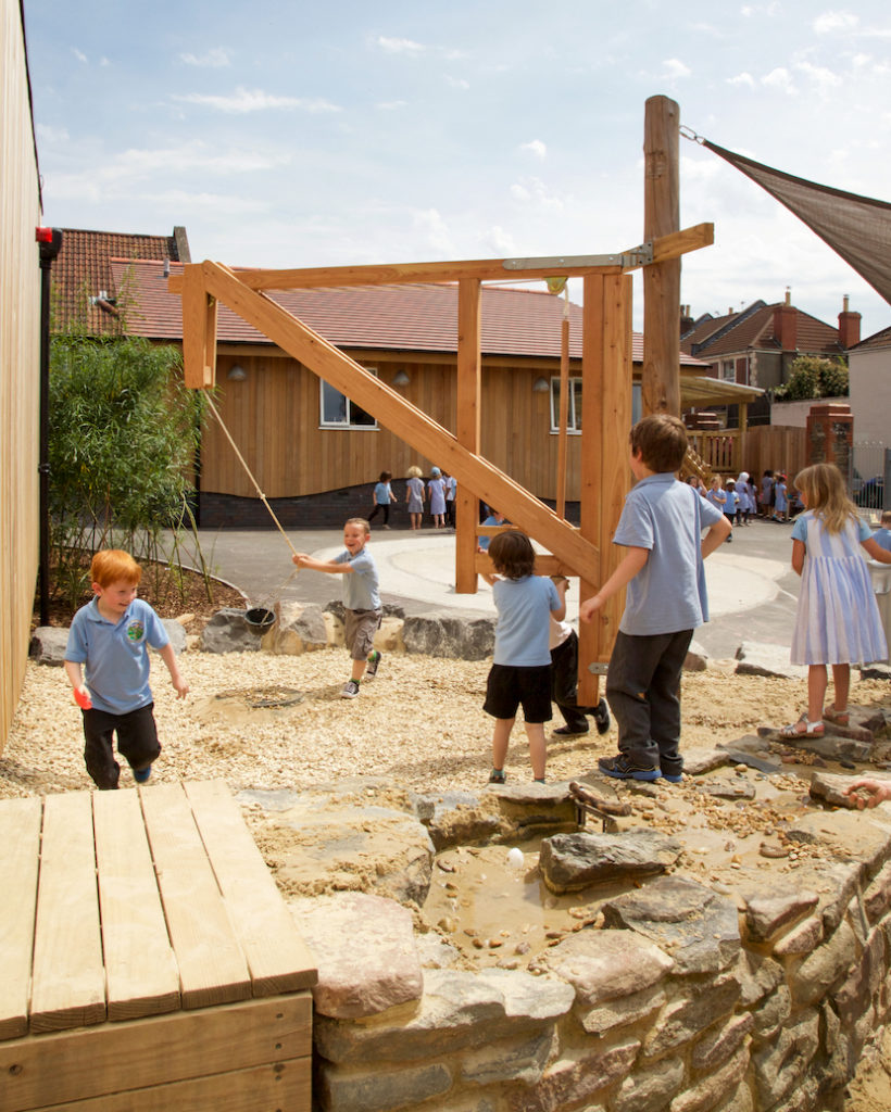 Children playing on a wooden playground equipment, a pulley with a bucket in an outdoor setting on a sunny day. A stone path leads up to the playground water play area and a shade sail is partially visible. A building with wood panelling is visible in the background.