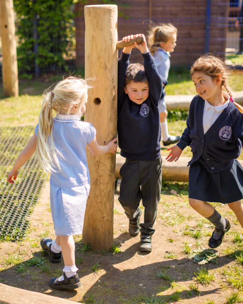 Three children in school uniforms interacting with a tall, cylindrical wooden den building pole play equipment with various holes drilled into it.