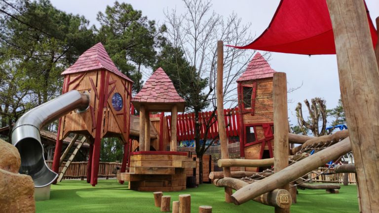 A large, intricate wooden playground set on bright green artificial turf, consisting of structures with pointed red roofs, a metal slide, and wooden logs and planks forming bridges and ladders. The playground is surrounded by trees and partially covered by a red shade canopy.