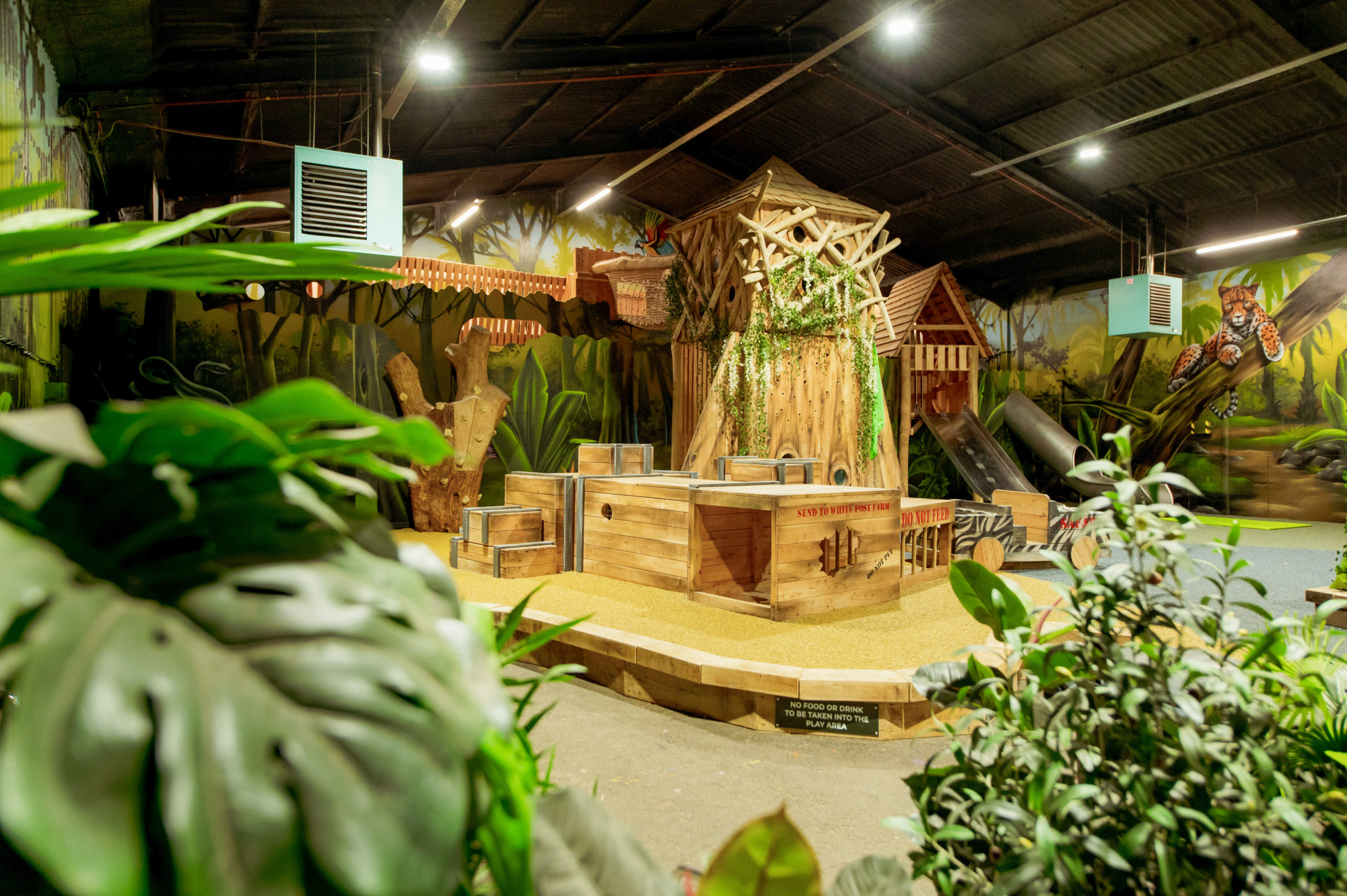 An indoor jungle-themed playground with various elements like bespoke wooden towers and structures, green plants, and painted walls.