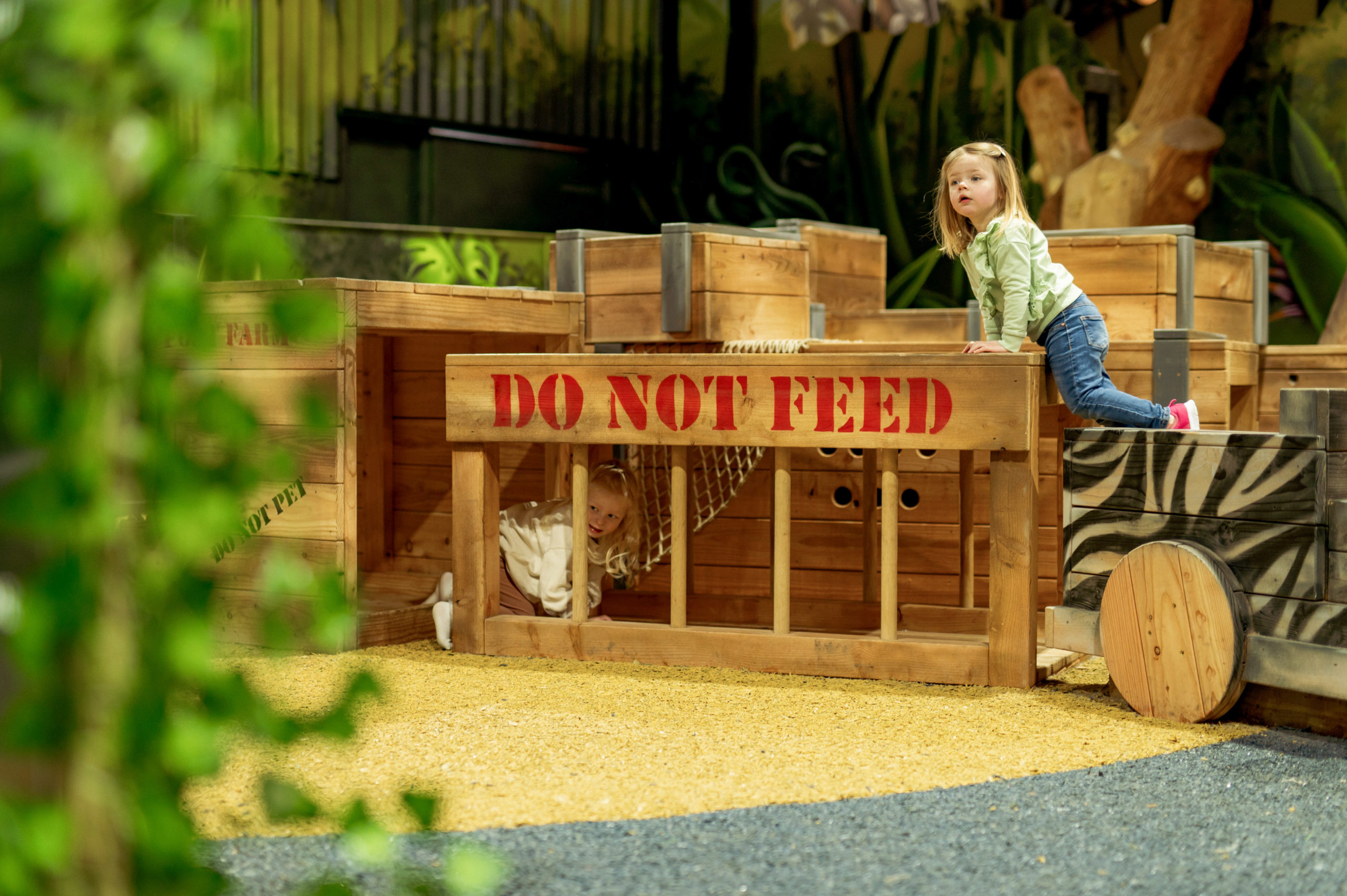 A playful scene where a child climbing on a wooden enclosure that has ‘DO NOT FEED’ written on it, while another child peeks out from inside the enclosure.
