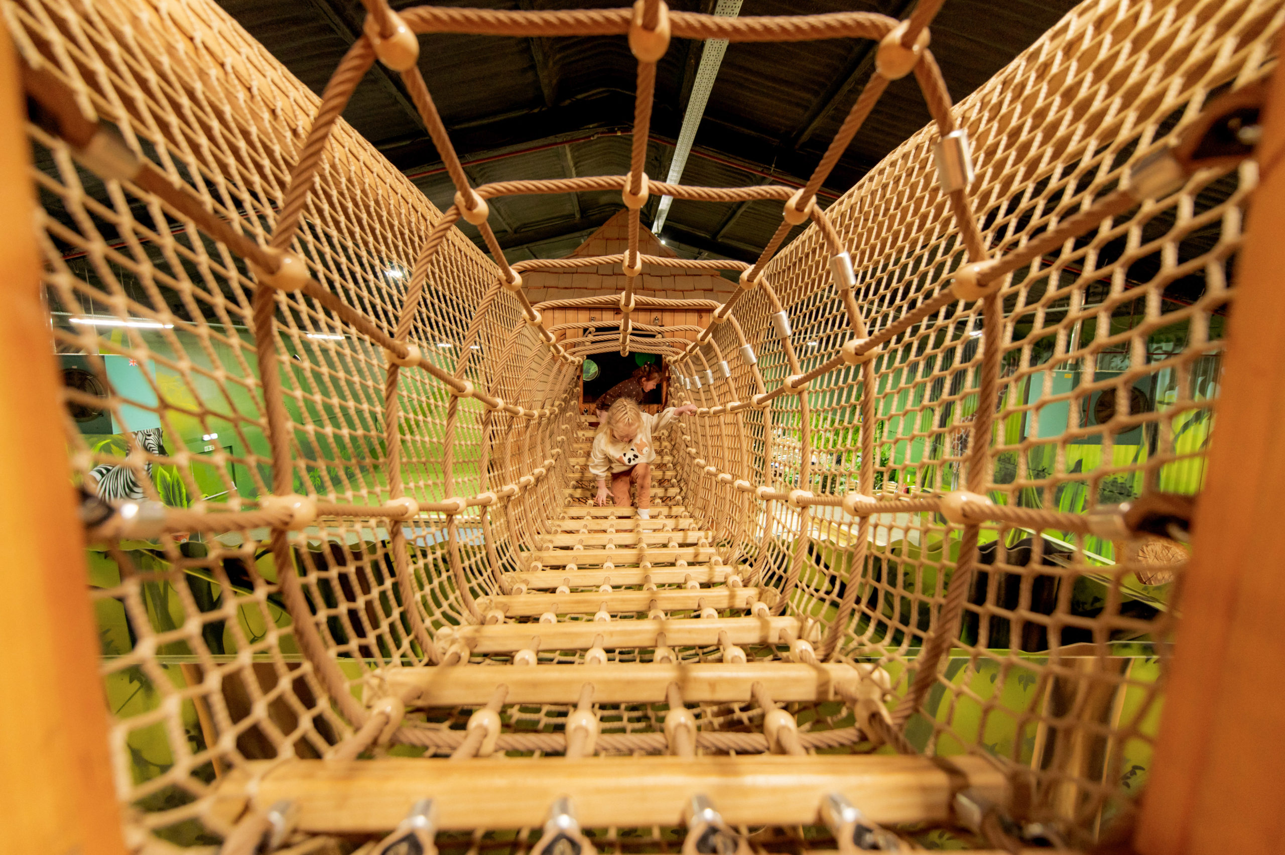 The interior view of a rope bridge inside a jungle-themed playground, showcasing its intricate woven structure and a child walking on it.