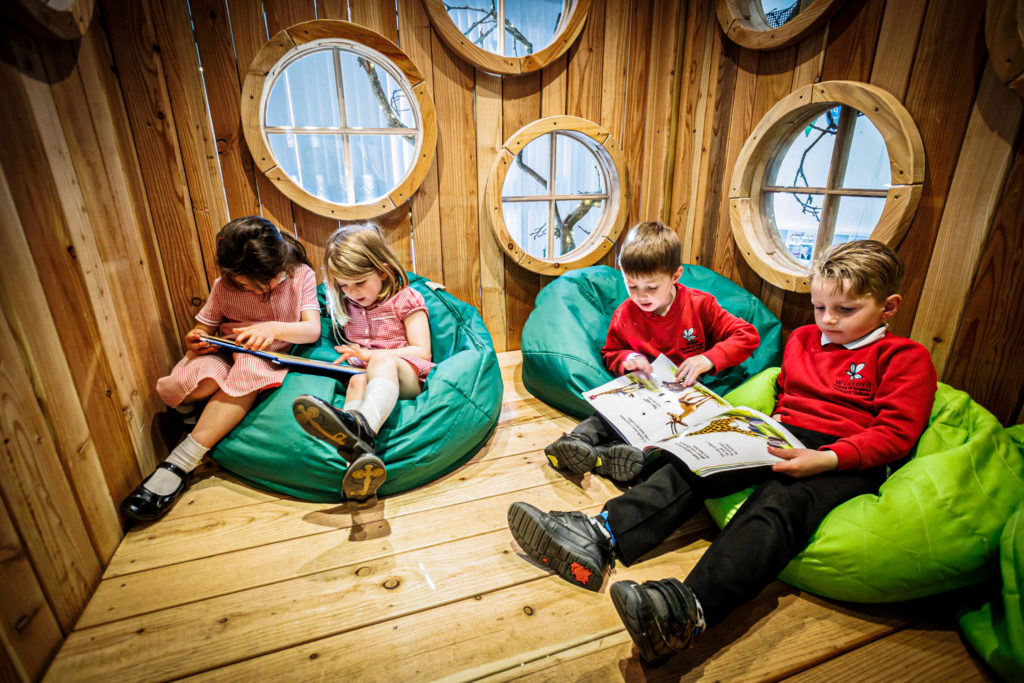 children are sitting in bean bags and reading books inside an indoor school library treehouse.