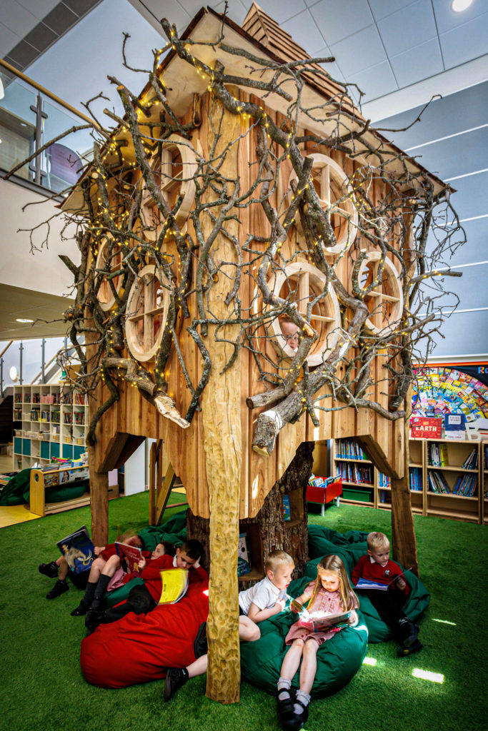 Children engrossed in reading books in a vibrant library, seated on colourful bean bags with a whimsical wooden treehouse as the centrepiece.