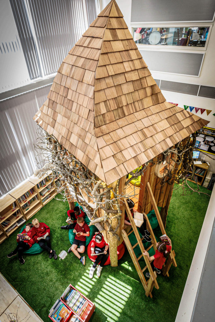 An indoor scene featuring a pyramid-shaped wooden structure built around a tree trunk, with children seated on red bean bags. The playful and vibrant atmosphere is enhanced by multicoloured triangular flags.