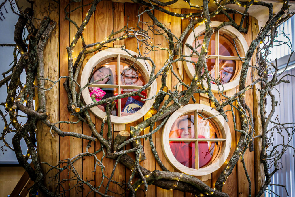 A whimsical wooden structure with three round windows, each with a person looking out. The structure is adorned with bare, leafless branches intertwined around it and fairy lights illuminating the scene.