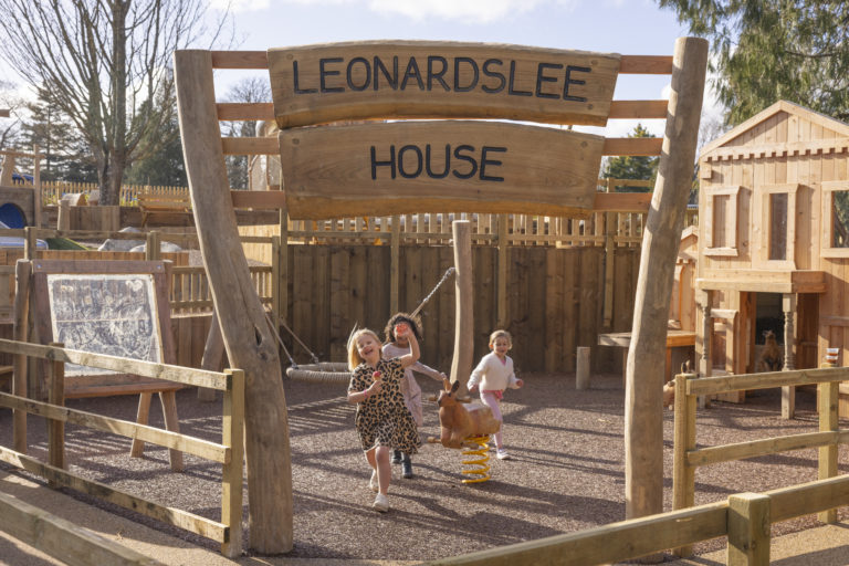 The entrance to Leonardslee House play area, with a wooden sign and two children running towards the gate on a sunny day.