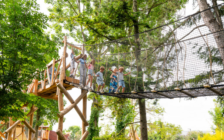 Group of individuals on an outdoor suspension bridge made of rope and wooden planks, surrounded by a safety net, in an adventure park or playground setting.